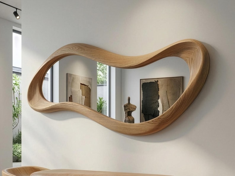 May include: A wall-mounted mirror featuring an irregular, wavy, organic-shaped frame crafted from light-toned wood with a visible grain pattern. The reflective surface displays a room interior with a framed painting and a small sculpture.