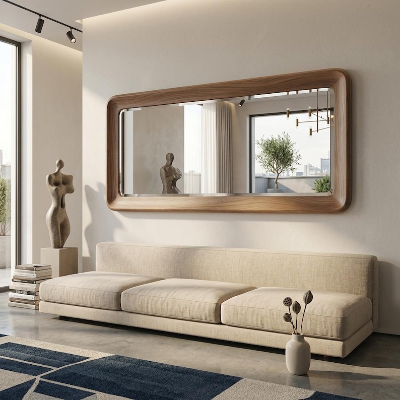 May include: A large rectangular mirror with a light brown wooden frame hangs above a beige sofa. The mirror reflects a room with a sculpture, a window, and a tree. A vase with dried flowers sits on the floor in front of the sofa.