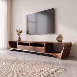 May include: A long, low-profile dark wood media console with curved, flowing edges and two central storage compartments. A flat-screen television is mounted on the wall above the console, which holds a small vase with dried grass and a glass vase.