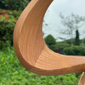 May include: A curved, light brown wooden frame with a visible natural grain pattern. The frame features a smooth, polished surface and a distinct, sweeping arc shape that connects to a reflective mirror surface, showcasing detailed wood joinery.