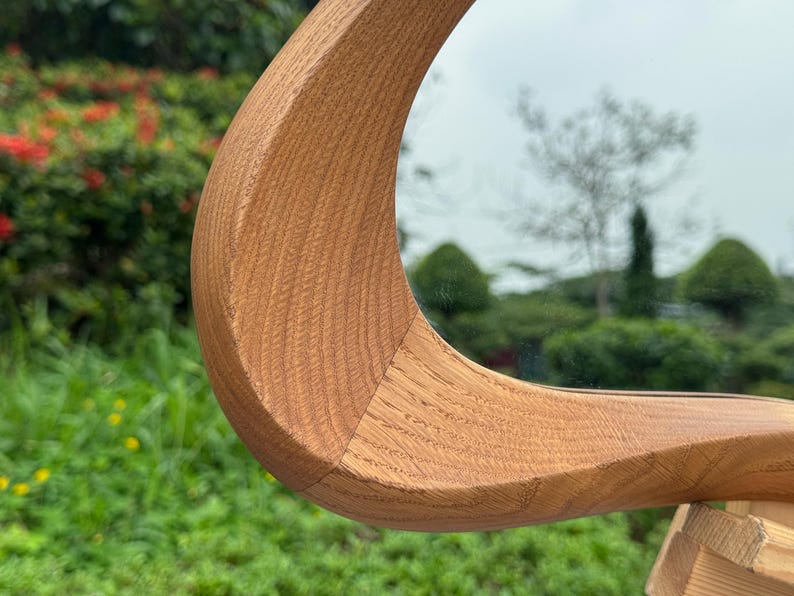 May include: A curved, light brown wooden frame with a visible natural grain pattern. The frame features a smooth, polished surface and a distinct, sweeping arc shape that connects to a reflective mirror surface, showcasing detailed wood joinery.