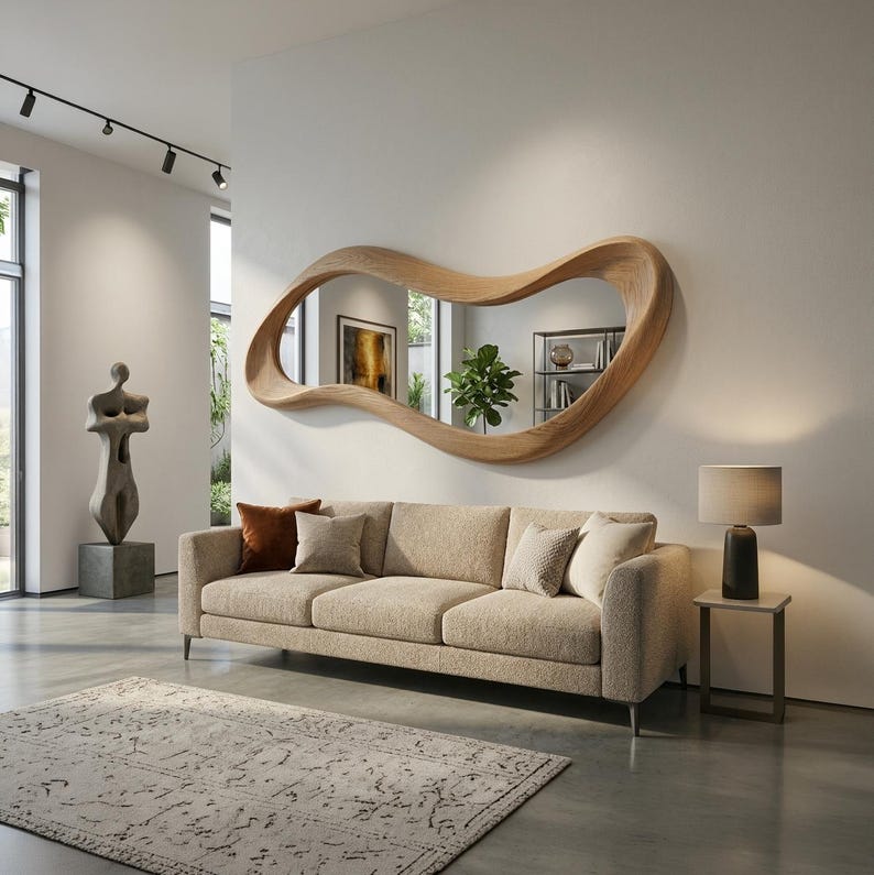 May include: A large, horizontal wall mirror featuring a thick, light-toned wooden frame with a fluid, organic, wavy shape. The mirror is mounted on a plain wall above a beige three-cushion sofa, reflecting a room interior with a green plant.