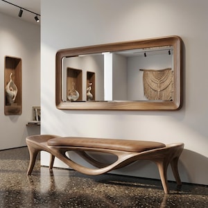 Op de afbeelding: Een moderne houten bank met een bruin leren zitting staat voor een grote rechthoekige spiegel met een houten frame. De spiegel weerspiegelt een kamer met decoratieve wandnissen en een macramé wandkleed. De bank heeft een uniek, gebogen ontwerp.