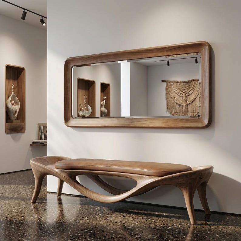 May include: A modern wooden bench with a brown leather seat is positioned in front of a large rectangular mirror with a wooden frame. The mirror reflects a room with decorative elements, including a macrame wall hanging.