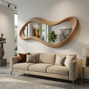 May include: A large, horizontal wall mirror featuring an irregular, organic, wavy wooden frame with a light brown wood grain finish. The mirror is mounted on a plain white wall above a beige textured sofa with several decorative throw pillows.