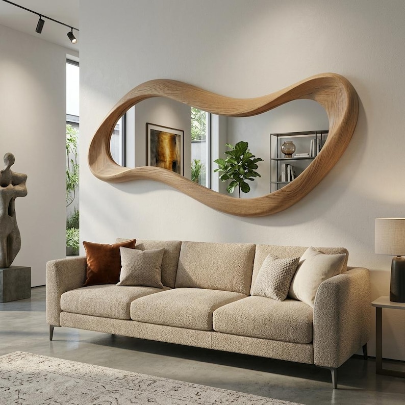 May include: A large, horizontal wall mirror featuring an irregular, organic, wavy wooden frame with a light brown wood grain finish. The mirror is mounted on a plain white wall above a beige textured sofa with several decorative throw pillows.