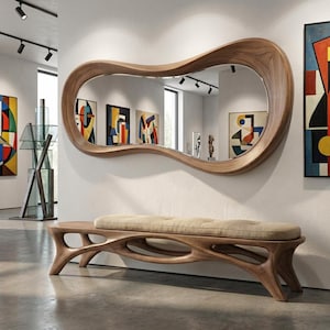 Op de afbeelding: Een grote, organisch gevormde houten spiegel met een walnootafwerking hangt boven een bijpassende bank met een gecapitonneerd, beige kussen. De bank heeft een uniek, open frame ontwerp. Kunstwerken sieren de muren.