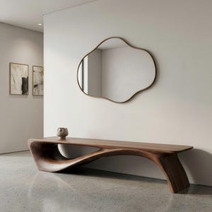 Puede incluir: Un banco de madera largo y de perfil bajo con una base curva continua y lisa y una superficie superior plana. Sobre el banco, un espejo de pared irregular en forma de nube con un marco metálico fino está montado en una pared clara.