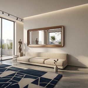 May include: A modern living room featuring a large rectangular mirror with a wooden frame above a beige sofa. A blue and white geometric rug covers the floor. A sculpture and vase with dried flowers add decorative elements.
