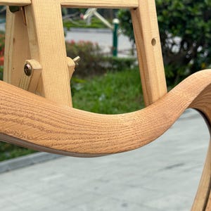 May include: A light-colored wooden mirror frame with a distinct wavy, organic shape. The wood grain is clearly visible across the curved surface. The mirror is mounted on a light-colored wooden easel stand with adjustable metal hardware components.