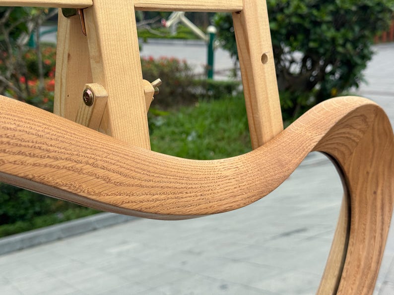 May include: A light-colored wooden mirror frame with a distinct wavy, organic shape. The wood grain is clearly visible across the curved surface. The mirror is mounted on a light-colored wooden easel stand with adjustable metal hardware components.