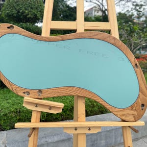 May include: An irregular, kidney-shaped mirror featuring a light-toned wooden frame with visible grain. The mirror surface has a smooth, light blue tint and includes several pre-drilled circular mounting holes along the perimeter of the wooden border.