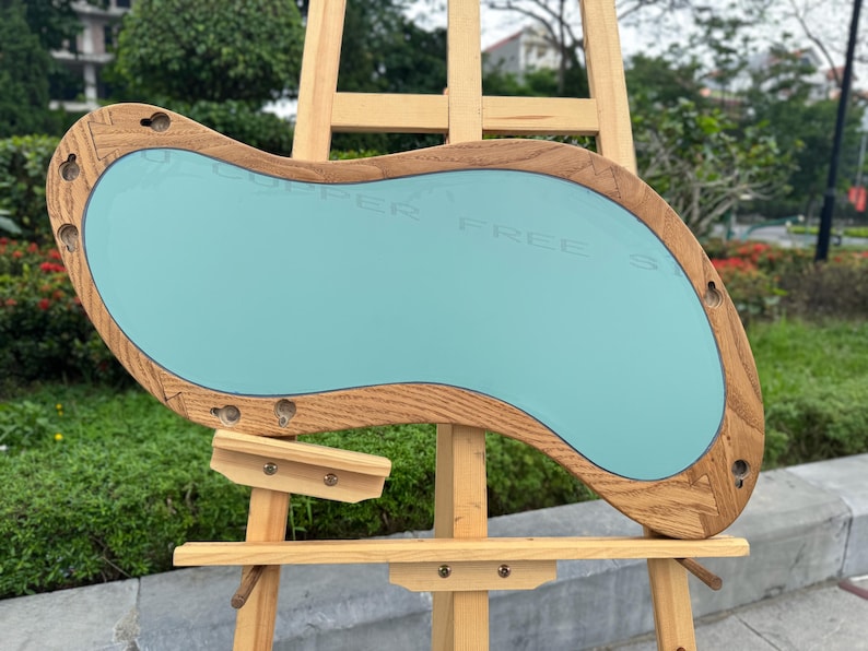 May include: An irregular, kidney-shaped mirror featuring a light-toned wooden frame with visible grain. The mirror surface has a smooth, light blue tint and includes several pre-drilled circular mounting holes along the perimeter of the wooden border.