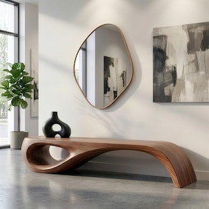 May include: A low-profile, curved wooden bench with a smooth, natural grain finish and a large, hollow oval cutout on one side. A black, abstract-shaped ceramic vase sits on the bench, and an irregular, organic-shaped wall mirror hangs above.