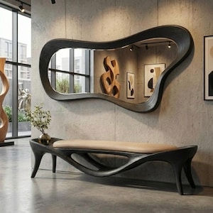 Op de afbeelding: Een zwarte houten bank met een gladde beige gestoffeerde zitting en een bijpassende zwarte houten wandspiegel met een onregelmatige, organische, vloeiende vorm. De bank heeft een gebogen, open onderstel en de spiegel reflecteert een kamer met kunstwerken.