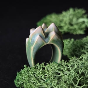 May include: A handcrafted ceramic ring with a mountain range design. The ring features a green and beige color scheme, with a textured surface. The ring is set against a dark background with green moss.
