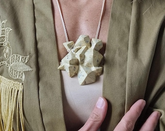 May include: A geometric pendant necklace with a light beige, textured, abstract design. The pendant hangs from a thin silver chain. The person is wearing a light brown jacket.