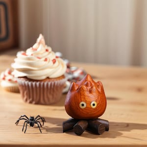 May include: A wooden sculpture of a cartoon fire character with yellow eyes, sitting on a wooden base. A black plastic spider is in front of the sculpture. Cupcakes with white frosting and red heart sprinkles are in the background.