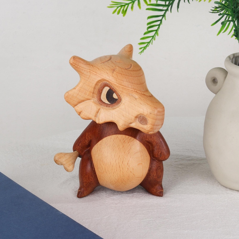 Puede incluir: Una figura de madera de Cubone, con una cabeza en forma de cr&aacute;neo marr&oacute;n claro y un cuerpo marr&oacute;n oscuro. La figura tiene una barriga redondeada y un peque&ntilde;o accesorio de hueso. Los ojos son oscuros, y el dise&ntilde;o general es detallado y liso.