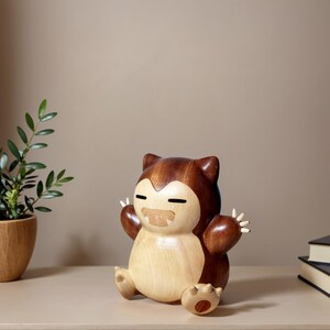 May include: A wooden sculpture of a cartoon character in a seated position with arms raised. The sculpture is made of light and dark brown wood, with a light-coloured belly and face. The character has closed eyes and a smiling mouth.