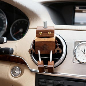 May include: A wooden robot car air vent clip. The robot has a star on its chest and is made of dark and light brown wood. It has a silver metal rod on top and gold-colored eye accents. The robot is sitting on a car air vent.