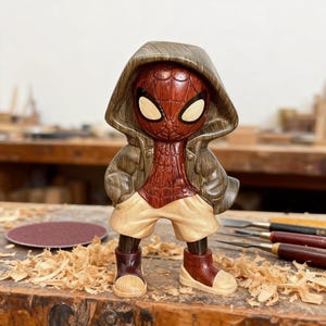 May include: A wooden Spider-Man figurine with a hooded jacket and shorts. The figure has a red and brown color scheme, with tan shorts and shoes. The figure is standing on a wooden surface with wood shavings.