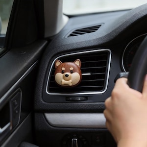 Könnte beinhalten: Ein braun-beiger, hundeförmiger Auto-Lufterfrischer ist an einer Lüftungsöffnung im Auto befestigt. Der Lufterfrischer hat ein knochenförmiges Detail und befindet sich vor einem schwarzen Armaturenbrett. Das Bild ist aus der Perspektive eines Beifahrers aufgenommen.