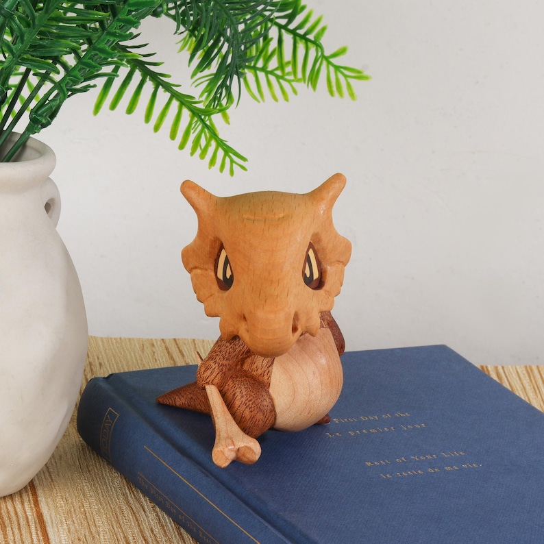 Puede incluir: Figura de madera hecha a mano de una criatura con cuerpo marr&oacute;n claro y detalles marr&oacute;n oscuro. La figura tiene ojos grandes y orejas puntiagudas. Est&aacute; sentada sobre un libro azul. Un jarr&oacute;n blanco con follaje verde est&aacute; en el fondo.