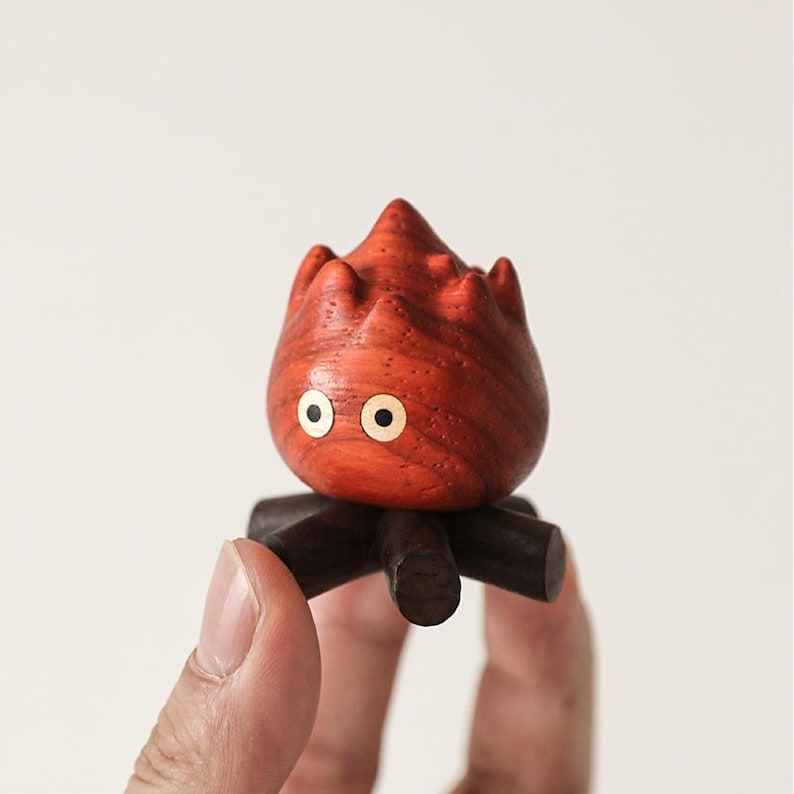 May include: A wooden figurine of a stylized flame with a red-orange finish and two round, white-rimmed eyes. The flame sits atop a dark brown, four-legged base, resembling a campfire. The figurine is held in a person's hand.