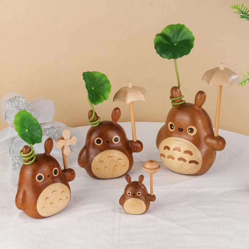 Pu&ograve; includere: Una collezione di statuette in legno raffiguranti il personaggio di Totoro. Le statuette marroni presentano dettagli in legno chiaro, occhi rotondi e motivi sul ventre. Alcune tengono piccoli ombrelli o foglie di loto. Diverse dimensioni sono presenti.