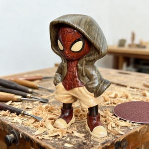 May include: A wooden carving of a superhero figure, featuring a red and black mask and costume, wearing a hooded jacket and shorts. The figure is posed with hands in pockets, and the carving is detailed with wood grain.
