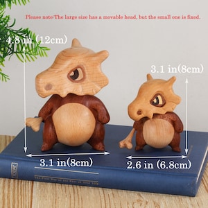 Puede incluir: Dos figuras de madera, una grande y otra peque&ntilde;a, que se asemejan a una criatura parecida a un dinosaurio. La figura m&aacute;s grande mide 12 cm de alto y 8 cm de ancho, mientras que la m&aacute;s peque&ntilde;a mide 8 cm de alto y 6,8 cm de ancho. El texto de la imagen dice "The First Day of the Rest of Your Life."
