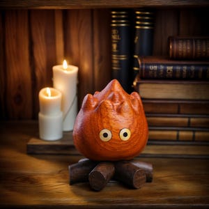 May include: A wooden sculpture of a fire demon with large, round eyes and a flame-like form. The sculpture is orange and sits on a wooden base. Two white candles are lit in the background, along with stacks of books.