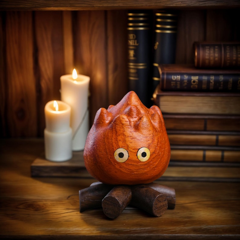 May include: A wooden sculpture of a fire demon with large, round eyes and a flame-like form. The sculpture is orange and sits on a wooden base. Two white candles are lit in the background, along with stacks of books.