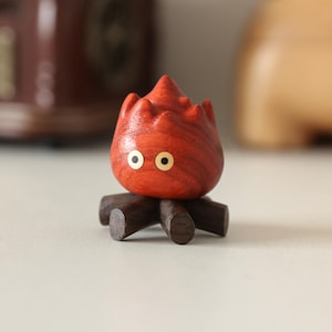 May include: A wooden sculpture of a fire spirit with a red-orange flame-like body and two golden eyes. The figure is mounted on a dark brown wooden base, resembling small logs. The handcrafted piece has a whimsical, cartoonish aesthetic.