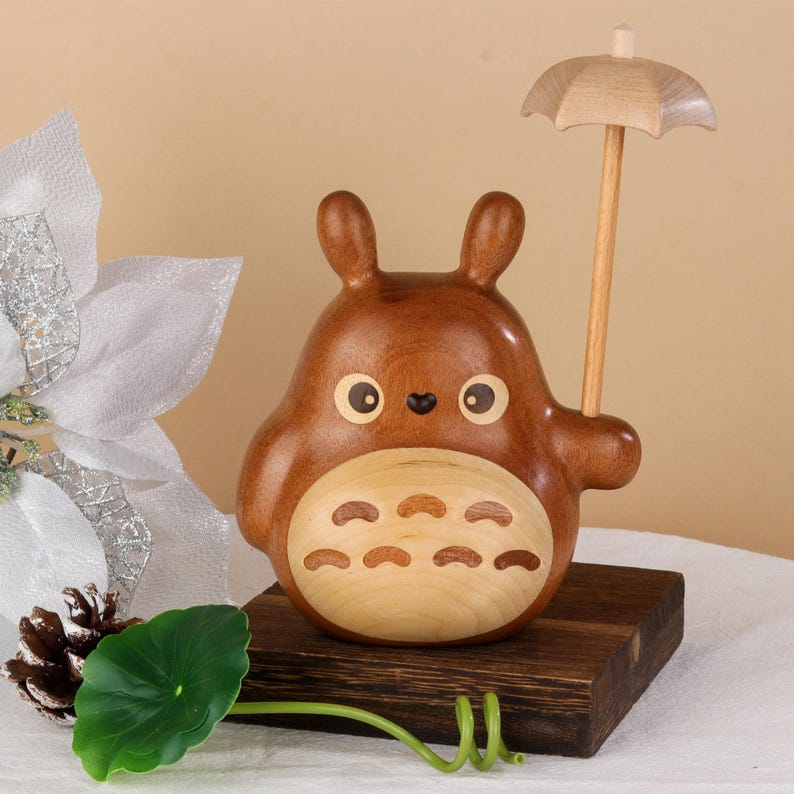 Pu&ograve; includere: Una statuetta in legno di un personaggio dei cartoni animati con corpo marrone, pancia chiara e grandi orecchie, che tiene un piccolo ombrello di legno. La statuetta &egrave; su una base di legno scuro. Gli occhi sono grandi e rotondi.