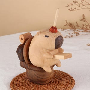 May include: A whimsical wooden capybara figurine seated on a toilet, holding a small wooden plank. The capybara has a plunger on its head. The toilet is made of dark wood, and the figurine sits on a woven mat.