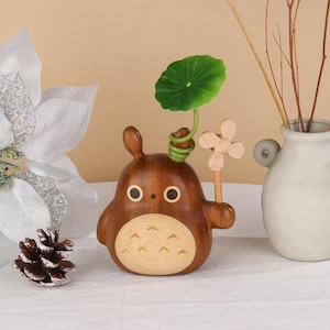 Pu&ograve; includere: Una statuetta in legno di un personaggio dei cartoni animati con un corpo rotondo, due piccole orecchie e due occhi circolari. Tiene in mano una piccola girandola di legno e ha una foglia verde e uno stelo attaccati alla testa. La statuetta &egrave; marrone con la pancia in legno pi&ugrave; chiaro.