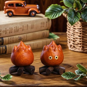 May include: Two wooden fire-shaped figurines with a dark brown base. One has two yellow eyes. An orange toy car and a stack of books are in the background. The figurines are on a wooden surface.