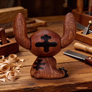 May include: A handcrafted wooden figurine of a cartoon character with large ears and a black emblem on its body. The figurine is made of dark brown wood and sits on a wooden surface. Other woodworking tools are visible in the background.