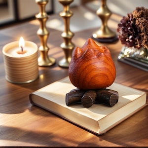 May include: A wooden sculpture resembling a flame, resting on a small wooden base, sits atop a book. A lit candle and gold candlesticks are in the background. The sculpture is a rich, reddish-brown color.