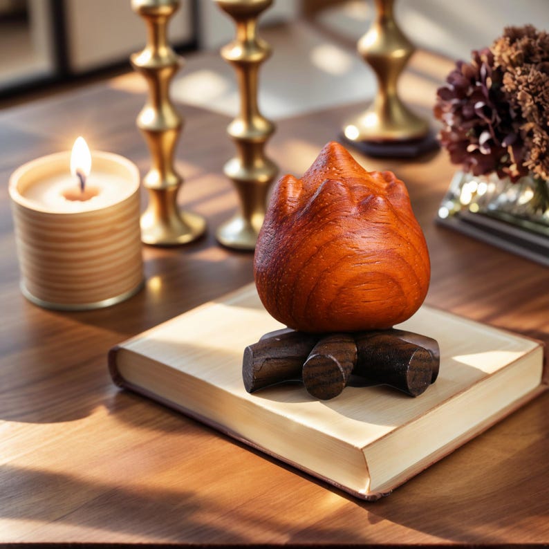 May include: A wooden sculpture resembling a flame, resting on a small wooden base, sits atop a book. A lit candle and gold candlesticks are in the background. The sculpture is a rich, reddish-brown color.