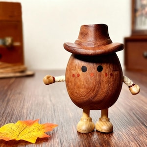 May include: A wooden figurine shaped like an egg, wearing a brown cowboy hat. The figurine has black eyes, red cheeks, and small wooden arms. It stands on wooden legs and feet. A fall leaf rests on the wooden surface.