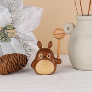 Pu&ograve; includere: Statuetta in legno di un personaggio dei cartoni animati con corpo marrone e pancia chiara, che tiene in mano un lecca lecca di legno. La statuetta ha grandi occhi rotondi e piccole orecchie. Una pigna di legno e un vaso bianco sono sullo sfondo.
