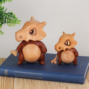 Puede incluir: Dos figuras de Cubone de madera, una grande y otra peque&ntilde;a, se exhiben sobre un libro azul. Las figuras tienen cabezas de color marr&oacute;n claro con forma de hueso y cuerpos de color marr&oacute;n oscuro. El libro tiene la palabra "TODAY" en el lateral.