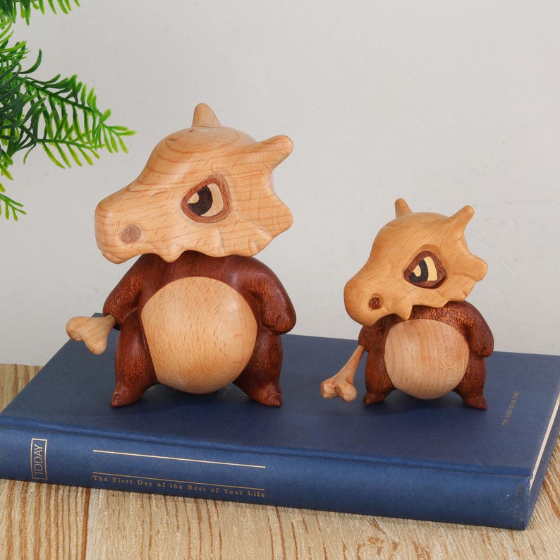 Puede incluir: Dos figuras de Cubone de madera, una grande y otra peque&ntilde;a, se exhiben sobre un libro azul. Las figuras tienen cabezas de color marr&oacute;n claro con forma de hueso y cuerpos de color marr&oacute;n oscuro. El libro tiene la palabra "TODAY" en el lateral.