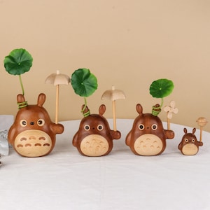 Pu&ograve; includere: Una collezione di quattro statuette in legno raffiguranti un personaggio dei cartoni animati. Le statuette sono marroni con pance in legno chiaro e grandi occhi. Alcune tengono piccoli ombrelli o accenti di foglie verdi, altre hanno un fiore o una lecca-lecca.
