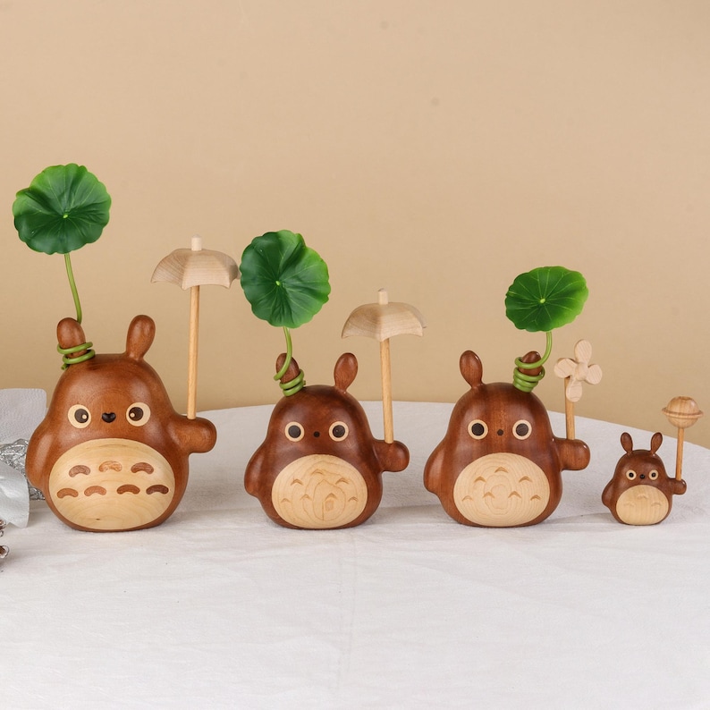 Pu&ograve; includere: Una collezione di quattro statuette in legno raffiguranti un personaggio dei cartoni animati. Le statuette sono marroni con pance in legno chiaro e grandi occhi. Alcune tengono piccoli ombrelli o accenti di foglie verdi, altre hanno un fiore o una lecca-lecca.