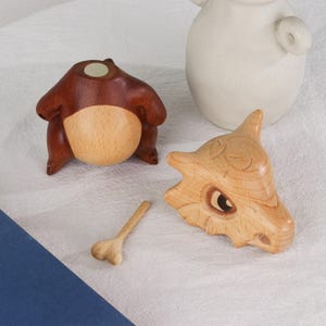 Puede incluir: Figuras de madera de una criatura parecida a un oso y una calavera, junto con un peque&ntilde;o hueso. La figura parecida a un oso es marr&oacute;n y beige, mientras que la calavera y el hueso son de madera clara. Las figuras son probablemente hechas a mano.