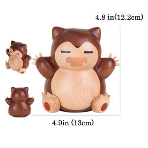 May include: Wooden Snorlax figure, approximately 12.2cm tall and 13cm wide. The figure is made of light and dark brown wood, with a smiling face and outstretched arms. Two smaller versions are also shown.